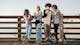 A group of four kids and children standing together. They wear sunglasses drink milkshakes and eat ice cream. All four have skateboards.